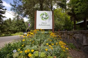 The Hoyt Arboretum Visitor Center Sign on Fairview Blvd near the Shuttle sign and with several bright yellow flowers in front.