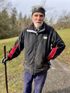 A man with grey hair and beard stands on a trail with one hand on his hip and another holding the handle of a rake. He is wearing jeans, a black rain coat spattered with mud, a black fleece headband, and work gloves.