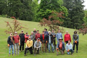 A group of about 20 volunteers gathered together on a grassy hillside in front of fruit trees with tall conifers in the background.