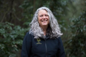 A woman with long, curly silver hair wearing a black fleece jacket with a green Hoyt Arboretum insignia on the right lapel smiles in the center of the frame surrounded by dark green foliage.