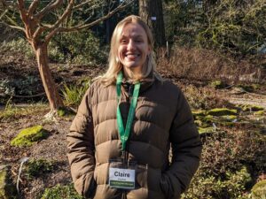 Blonde female from the waist up wearing an army green puffy coat and a lanyard with her name Claire on it. She is standing in from of a rock garden with a bare branched tree.